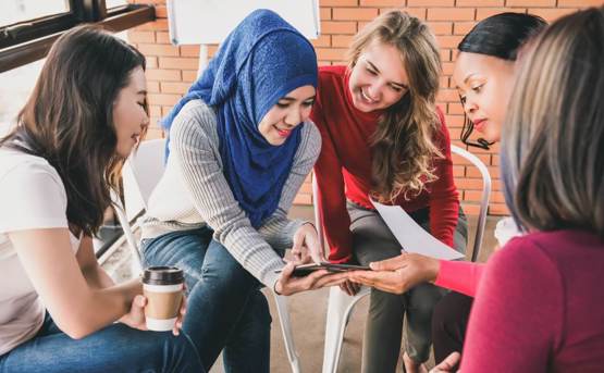 young women of different ethnicity crowd round a tablet smiling 